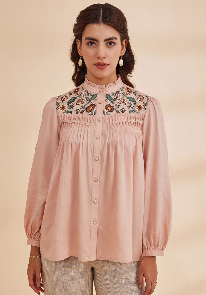 A woman wearing a pink top with floral embroidery at the yoke, full sleeves, and a round neckline.
